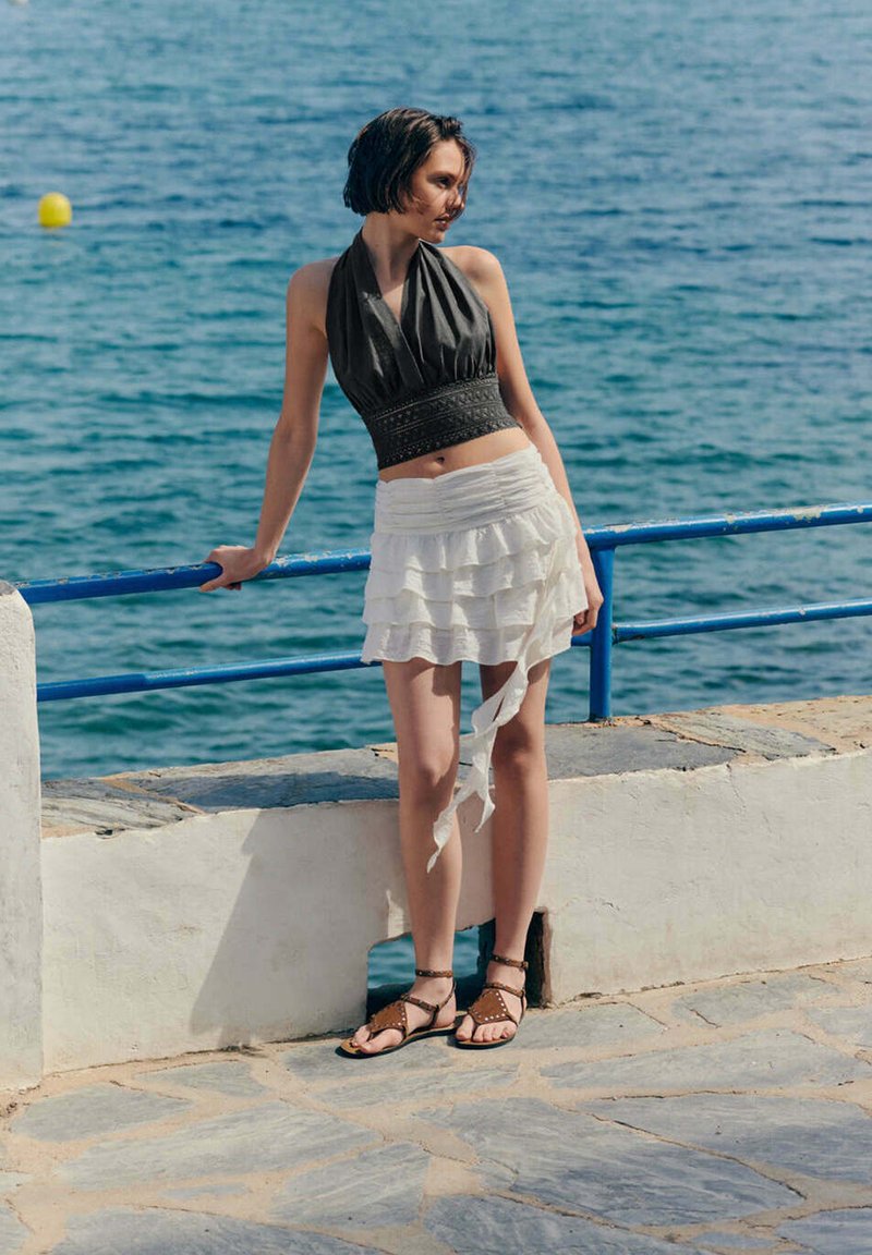 Jeune femme en haut noir sans manches et jupe blanche à volants s’appuie sur une rambarde bleue près de la mer sur une promenade pavée ensoleillée.