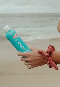 Turkusowa butelka sprayu przeciwsłonecznego z białą nakrętką, zdobiona różowymi kwiatowymi akcentami i etykietą SPF "50". Ręka z czerwonym aksamitnym gumką do włosów.