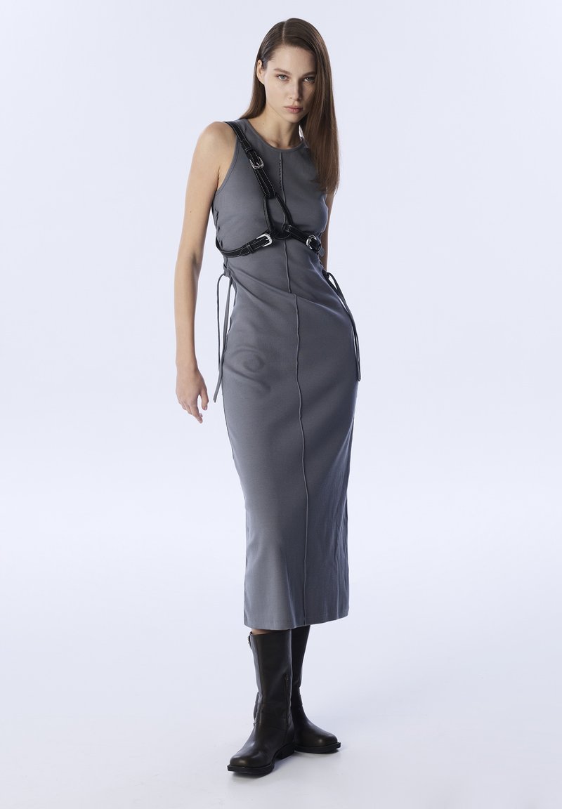 Vestido gris sin mangas con una silueta ajustada, que presenta tirantes de arnés negros en contraste, lazos laterales y botas negras hasta la rodilla.