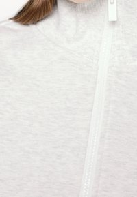 Gros plan d’un pull blanc à fermeture éclair avec col côtelé et longs cheveux bruns sur le côté gauche.