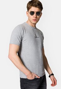 Szara koszulka z okrągłym dekoltem i krótkim rękawem, z małym czarnym logo, w zestawie z czarnymi dżinsami i okrągłymi okularami przeciwsłonecznymi.