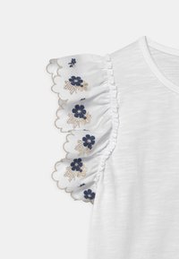 Biała koszulka z falbanami na rękawach ozdobionych haftem w niebieskie kwiaty i złotymi akcentami, na jednolitym tle.