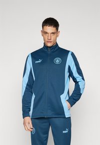 PUMA športna jakna v temno modri barvi s svetlo modrimi stranskimi paneli. Ima zadrgo spredaj, visok ovratnik in logo Manchester City na prsih.