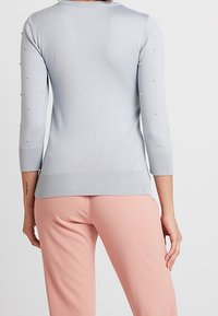 Ljusblå långärmad tröja med små strukturerade prickar på ärmarna, bärs med ljusrosa byxor, visad bakifrån.