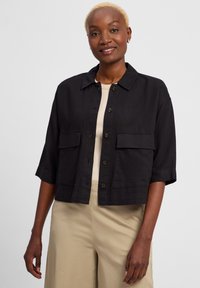 Zwarte cropped jas van een gestructureerde stof, met grote borstzakken en bruine knopen. Gedragen over een beige top en gecombineerd met lichte pants.