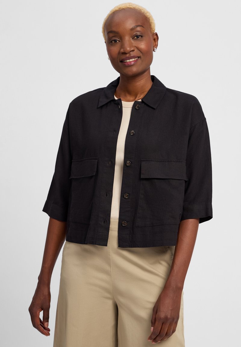 Zwarte cropped jas van een gestructureerde stof, met grote borstzakken en bruine knopen. Gedragen over een beige top en gecombineerd met lichte pants.