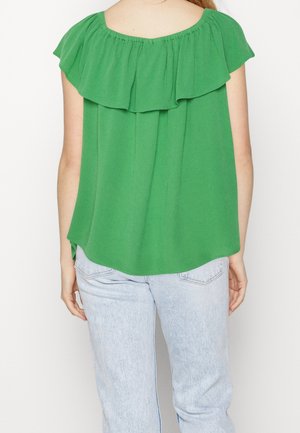 Kvinde iført en grøn flæset off-shoulder top og lyseblå jeans, vist bagfra mod en ensfarvet hvid baggrund.