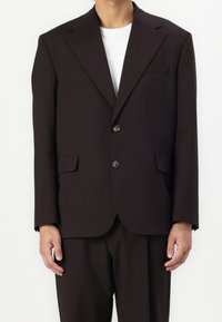 Ruskea räätälöity bleiseri, jossa on viiltohuppu, kaksi etutaskua, napin suljenta ja se on yhdistetty valkoiseen pyöreäkaulaiseen paitaan.