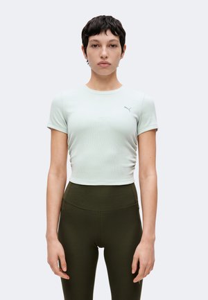 Femme aux cheveux courts et foncés portant un haut ajusté à manches courtes vert clair et des leggings taille haute vert foncé, debout devant un fond uni.