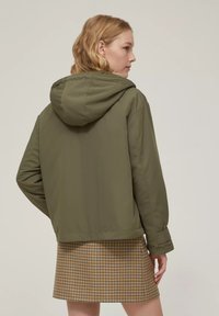 Giacca a vento verde oliva con cappuccio e vestibilità rilassata, indossata sopra una gonna a quadri. Tessuto liscio con finitura opaca.