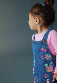 Salopette in denim con ricami colorati che includono fiori, frutti e stelle. Indossata sopra una maglia rosa a maniche lunghe. Caratterizzata da bottoni rotondi marroni.