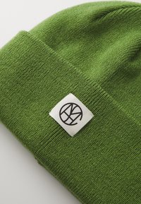 Green knitted beanie with a soft texture. Features a white label sewn on, displaying a black circular logo with stylized letters.