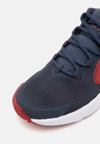 Chaussure de running marine avec un dessus texturé à rayures verticales, des accents rouges sur la semelle et une semelle intermédiaire blanche.