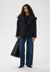 Marineblauer Mantel aus Wollmischung mit weitem Kragen und doppelreihigen Knöpfen. Kombiniert mit ausgewaschenen blauen Jeans mit weitem Bein und schwarzen Accessoires.