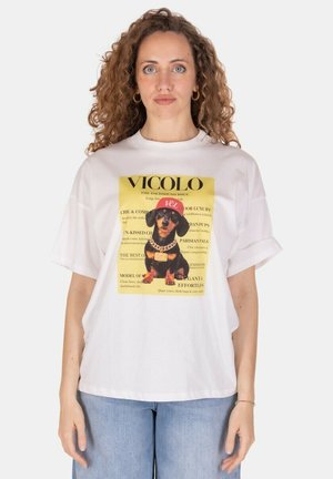 Donna con capelli ricci indossa una t-shirt bianca con un bassotto con cappellino rosso e catena dorata su sfondo giallo, abbinata a jeans azzurri.