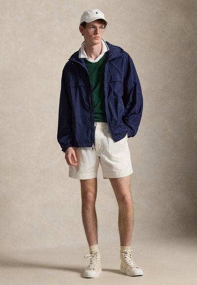 Jeune homme portant des lunettes rondes, une casquette blanche, une veste bleu marine, un pull vert, un polo blanc, un short blanc et des baskets montantes beiges, debout devant un fond beige.