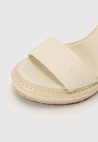 Beige glidesandal med en bred, preget stropp, teksturert espadrille-såle og en sydd juteakutt rundt kanten.