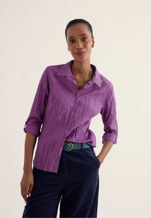 Femme portant une chemise violette froissée à boutons avec manches retroussées, pantalon sombre et ceinture turquoise, debout avec une main dans la poche.
