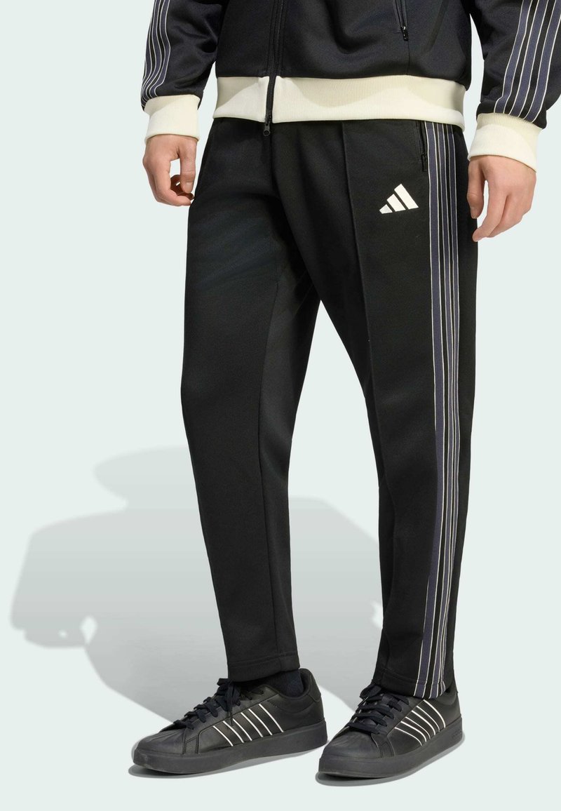 Homme portant un pantalon de survêtement Adidas noir avec des bandes blanches latérales et un logo, associé à des baskets noires à bandes blanches.