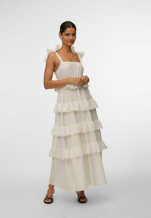 Kvinde iført en hvid, ærmeløs lagdelt maxikjole med båndstropper og flæsedetaljer, stående med sammenfoldede hænder og sorte sandaler med stropper.