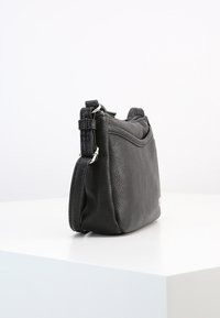 Černá texturovaná kožená crossbody taška s nastavitelným popruhem a stříbrným kovovým hardwarovým příslušenstvím, vystavená na bílé podložce proti jednoduchému pozadí.