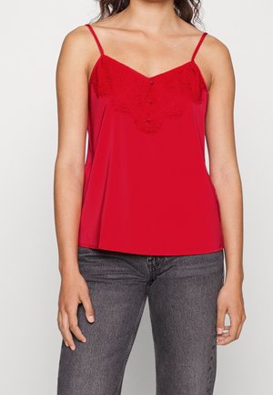 Kobieta ubrana w czerwoną, bezrękawną koszulkę na ramiączkach z koronkowym wykończeniem i guzikami, w połączeniu z ciemnoszarymi dżinsami.