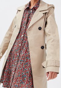 Woman wearing a beige trench coat over a red floral button-up dress, with black buttons and wrist strap details on the coat.