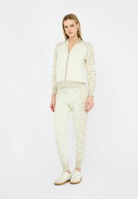 Giacca beige chiaro con zip e maniche con stampa animalier, abbinata a pantaloni affusolati coordinati. Entrambi presentano polsini a costine e una texture morbida.