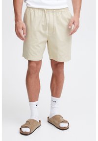 Light beige cotton shorts with an elastic waistband and adjustable drawstring. Worn with white socks and brown sandals.