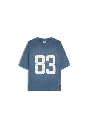T-shirt bleu à manches courtes avec un grand numéro blanc 83 à l'avant et une petite étiquette rouge près de l'ourlet inférieur du côté gauche.