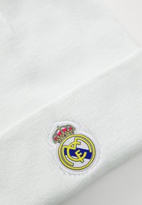 White knit beanie featuring an embroidered Real Madrid logo with a crown, blue and yellow accents, and a textured finish.