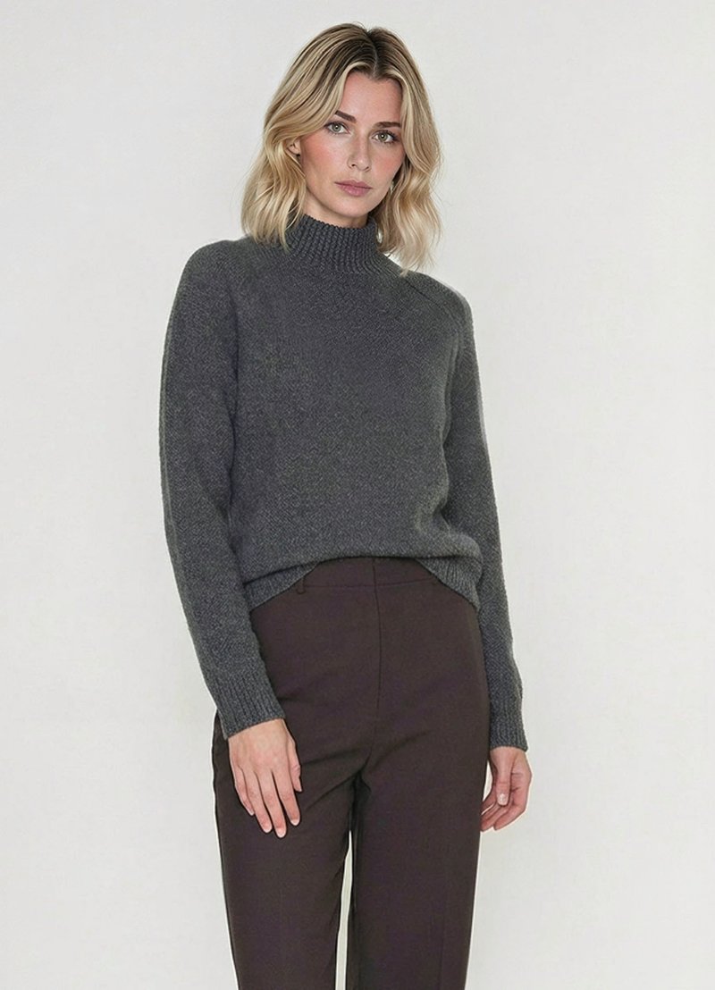 Donna con capelli biondi che indossa un maglione a collo alto grigio scuro e pantaloni marroni scuri, in piedi davanti a uno sfondo chiaro e uniforme.