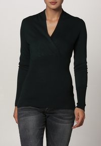 Mørkegrøn strikket sweater med lange ærmer, V-hals og ribbet tekstur, der har en tætsiddende silhuet og lagdelt design ved brystet.