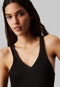 Top in tessuto a costine nere con scollatura a V e spalline larghe. Il tessuto ha una texture liscia e presenta un design pulito e minimalista.