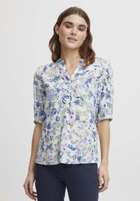 Blouse à imprimé floral avec un col en V, présentant des motifs bleu, vert et lavande sur un fond clair ; coupe ample et manches trois-quarts.