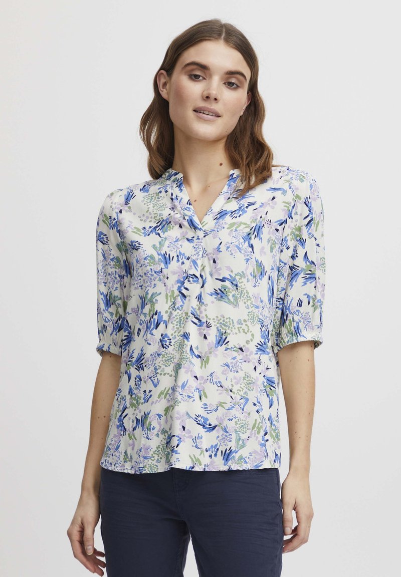 Blouse à imprimé floral avec un col en V, présentant des motifs bleu, vert et lavande sur un fond clair ; coupe ample et manches trois-quarts.