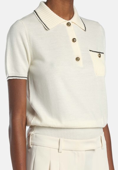 Kremowy pulower z krótkim rękawem, z prążkowanym kołnierzem, zapięciem na trzy guziki z przodu, naszywaną kieszenią oraz czarnym przeszyciem na krawędziach kołnierza i rękawów.