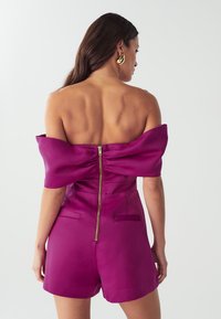 Romper fucsia scollato con zip sul retro, maniche strutturate e due piccole tasche. Texture satinata liscia.