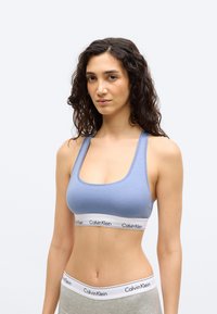 Γυναίκα με σκουρόχρωμα σγουρά μαλλιά που φοράει ανοιχτό γαλάζιο αθλητικό σουτιέν Calvin Klein και γκρι εσώρουχο Calvin Klein μπροστά σε απλό φόντο.