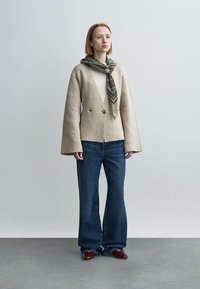 Μπεζ μπουφάν τύπου wrap με μανίκια καμπάνα, δύο κουμπιά, συνδυασμένο με σκούρο μπλε τζιν φαρδιά στο κάτω μέρος και πράσινο φουλάρι με σχέδια δεμένο στο λαιμό.