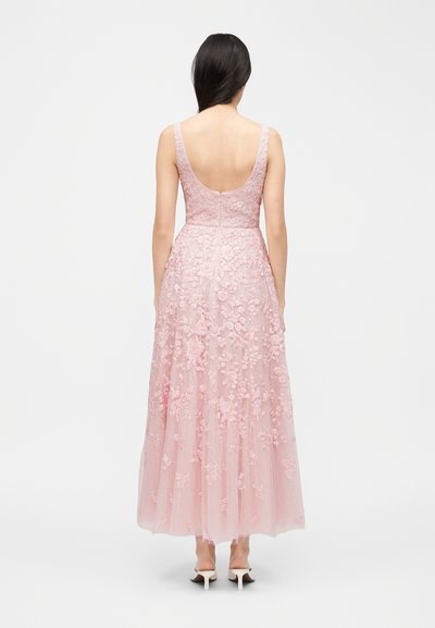 Γυναίκα με μακριά σκουρόχρωμα μαλλιά, που φοράει ένα αμάνικο ροζ φόρεμα δαντέλας μέχρι το πάτωμα με κεντήματα λουλουδιών, φαίνεται από την πλάτη σε λευκό φόντο.