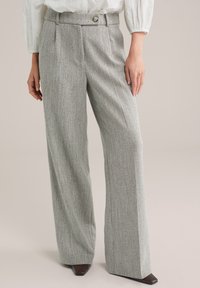 Femeie purtând pantaloni gri din țesătură herringbone, cu croială largă pe picior și talie cu nasturi, asortați cu pantofi maro cu vârf pătrat.