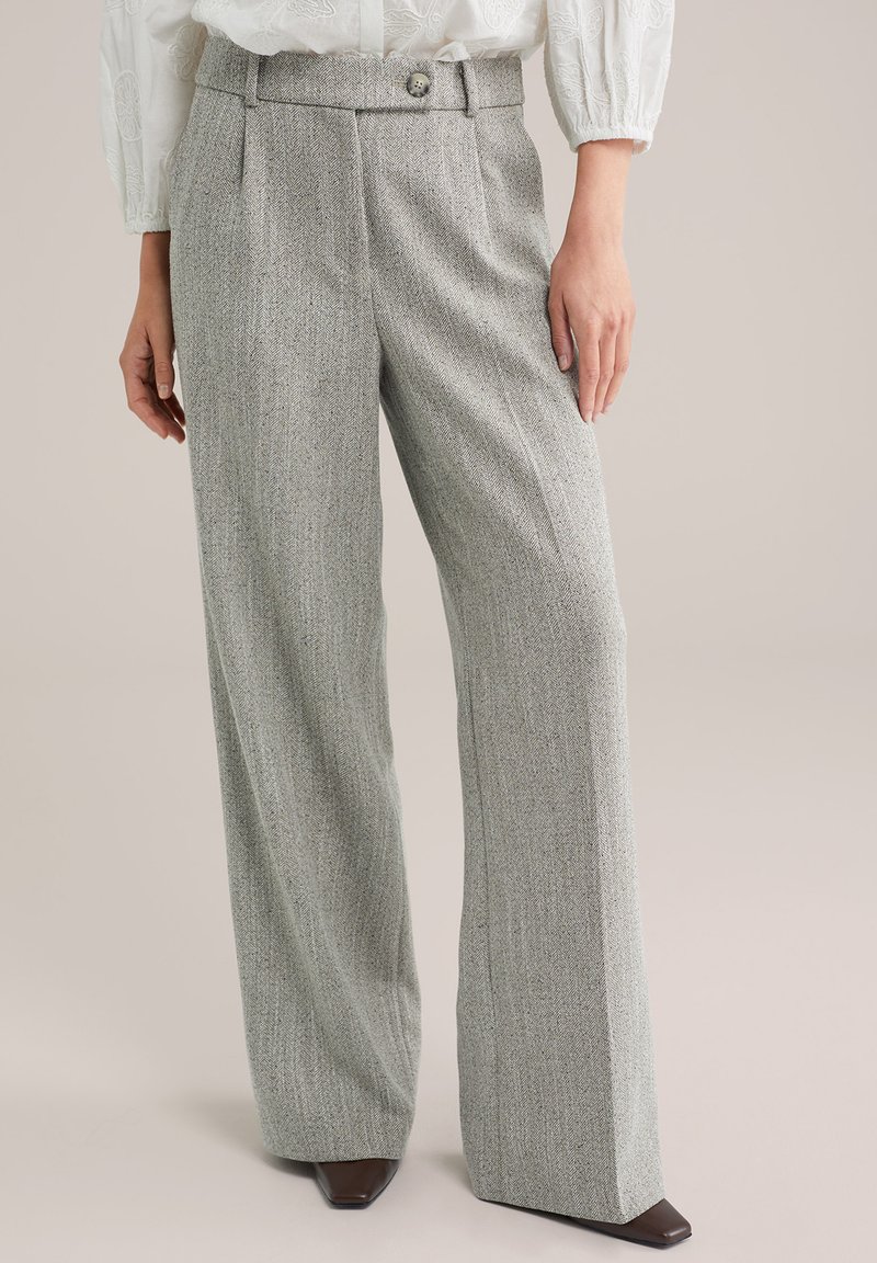 Femeie purtând pantaloni gri din țesătură herringbone, cu croială largă pe picior și talie cu nasturi, asortați cu pantofi maro cu vârf pătrat.