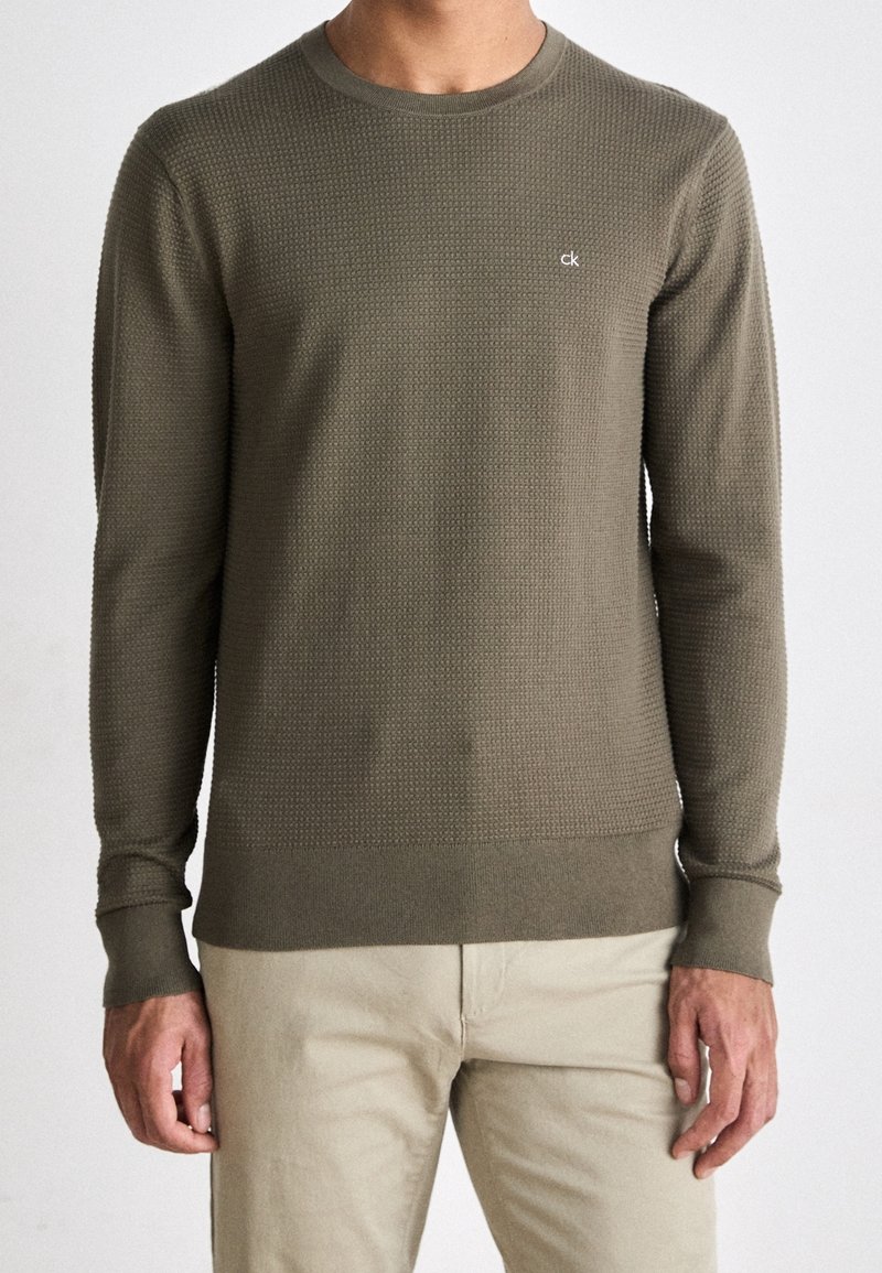 Olivgrüner, langärmeliger Strickpullover mit strukturiertem Muster, Rundhalsausschnitt sowie gerippten Bündchen und Saum. Verfügt über kleines Logo auf der Brust.