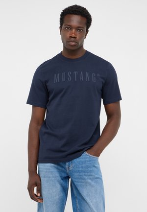 Junger Mann mit kurzen lockigen Haaren, der ein marineblaues Mustang-T-Shirt und hellblaue Jeans trägt und mit einer Hand in der Tasche steht.