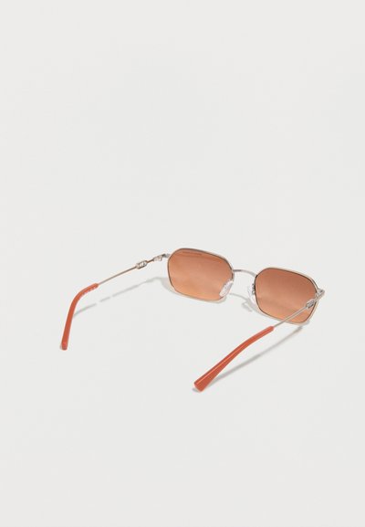 Lunettes de soleil rectangulaires avec des montures fines en métal argenté, des verres teintés orange-brun et des branches avec embouts en plastique orange, posées sur une surface blanche.