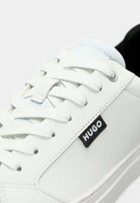 Λευκό αθλητικό παπούτσι με λείο συνθετικό επάνω μέρος, επίπεδα κορδόνια, ασημί ροδέλες και μαύρο ταμπελάκι με λογότυπο στο πλάι. Στρογγυλεμένη μύτη και μινιμαλιστικός σχεδιασμός.