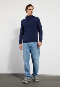 Marinblå stickad hoodie med krage och dragsko, matchad med ljusblå lösa jeans och grå sportskor. Slät textur och avslappnad passform.