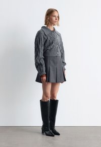 Grauer Kabelstrickpullover mit großem Kragen und Knöpfen, kombiniert mit einem gefalteten grauen Rock. Schwarze, knielange, glänzende Stiefel mit Krokodilprägung.