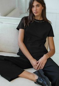 Haut noir à manches courtes avec un accent de chaîne autour du col, associé à un pantalon noir ample et des chaussures noires, assis sur un canapé clair.